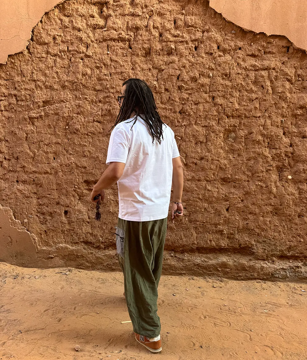 Desert house wall with exposed mud bricks. A man with dreads is walking with his back turned.